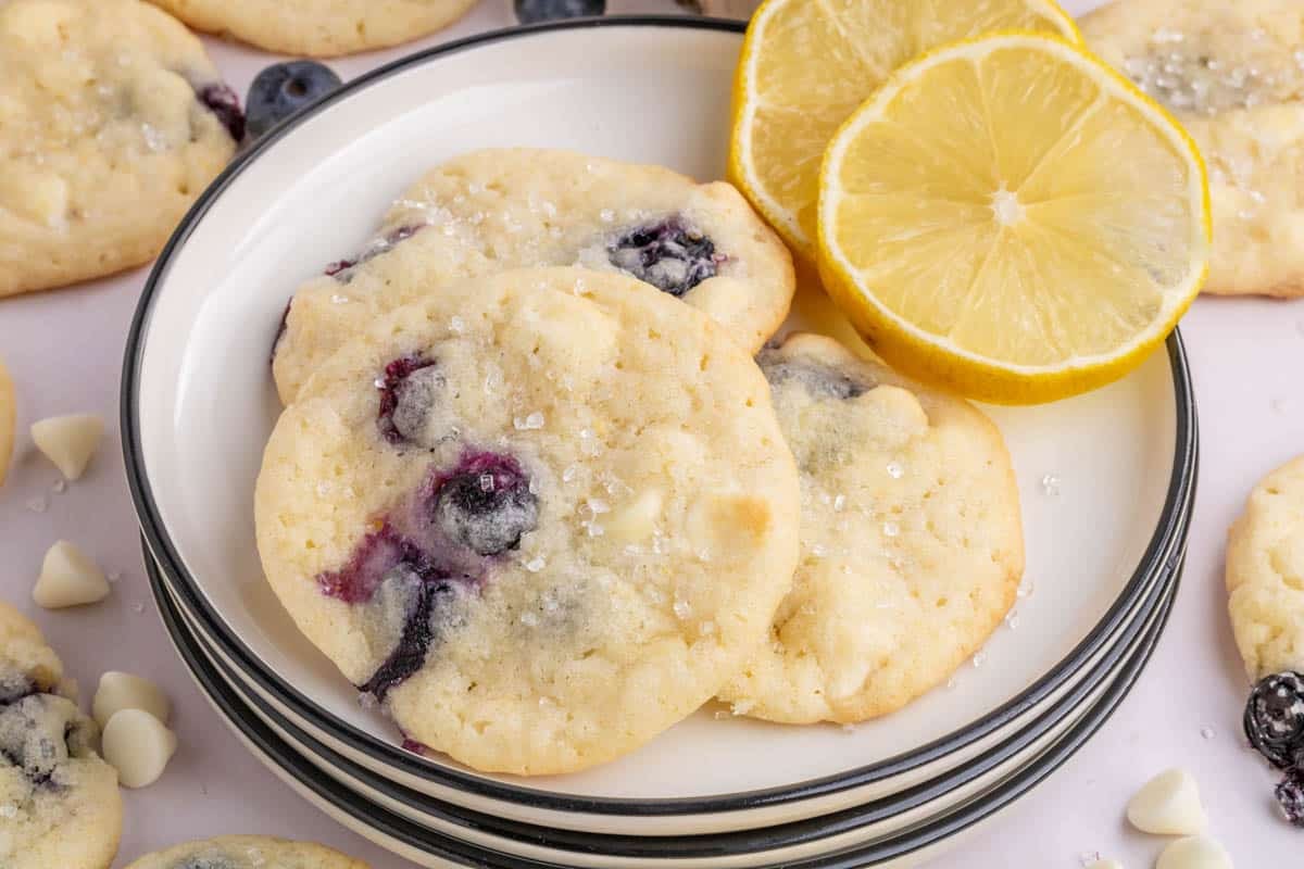 A plate of blueberry and white chocolate cookies is shown with two lemon slices, surrounded by more cookies and white chocolate chips.