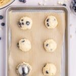 Six scoops of blueberry lemon cookie dough on a parchment-lined baking sheet, with a cookie scoop, blueberries, white chocolate chips, and lemon visible around the tray.