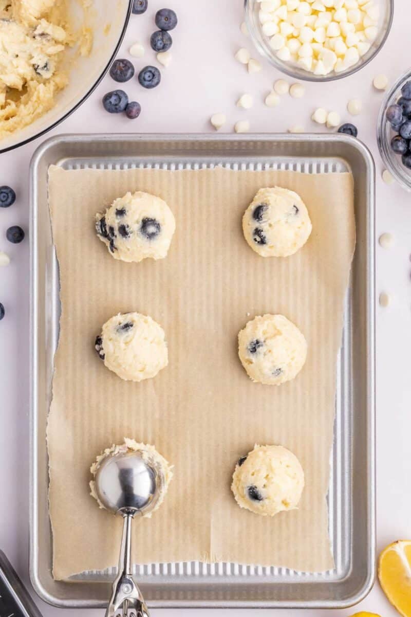 Six scoops of blueberry lemon cookie dough on a parchment-lined baking sheet, with a cookie scoop, blueberries, white chocolate chips, and lemon visible around the tray.