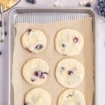 Six lemon blueberry cookies on a parchment-lined baking sheet, surrounded by bowls of cookie dough, sugar, white chocolate chips, and fresh blueberries.