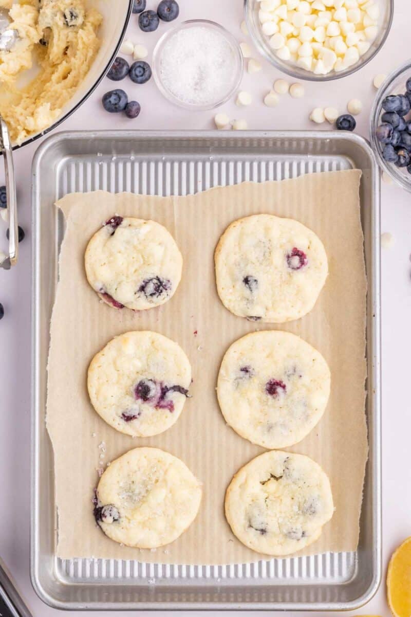 Six lemon blueberry cookies on a parchment-lined baking sheet, surrounded by bowls of cookie dough, sugar, white chocolate chips, and fresh blueberries.