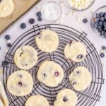 Nine lemon blueberry cookies cool on a wire rack, with bowls of white chocolate chips, blueberries, and sugar nearby; lemons and a baking sheet are also visible.