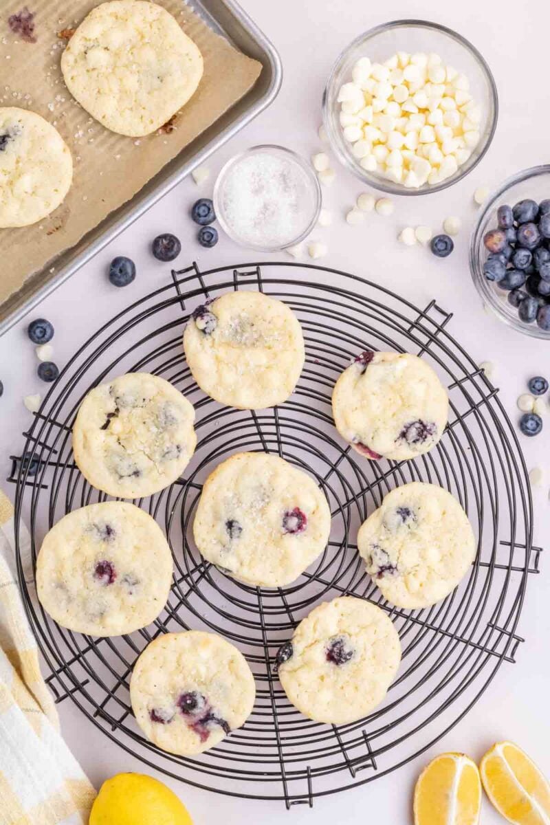 Nine lemon blueberry cookies cool on a wire rack, with bowls of white chocolate chips, blueberries, and sugar nearby; lemons and a baking sheet are also visible.