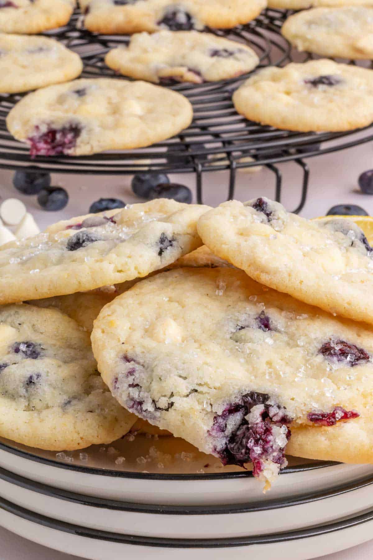 A plate of blueberry lemon cookies with visible sugar crystals, with more cookies cooling on a wire rack in the background.