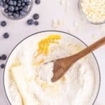 A mixing bowl with flour, butter, egg, and a wooden spoon, surrounded by bowls of blueberries, white chocolate chips, vanilla extract, and a lemon wedge.