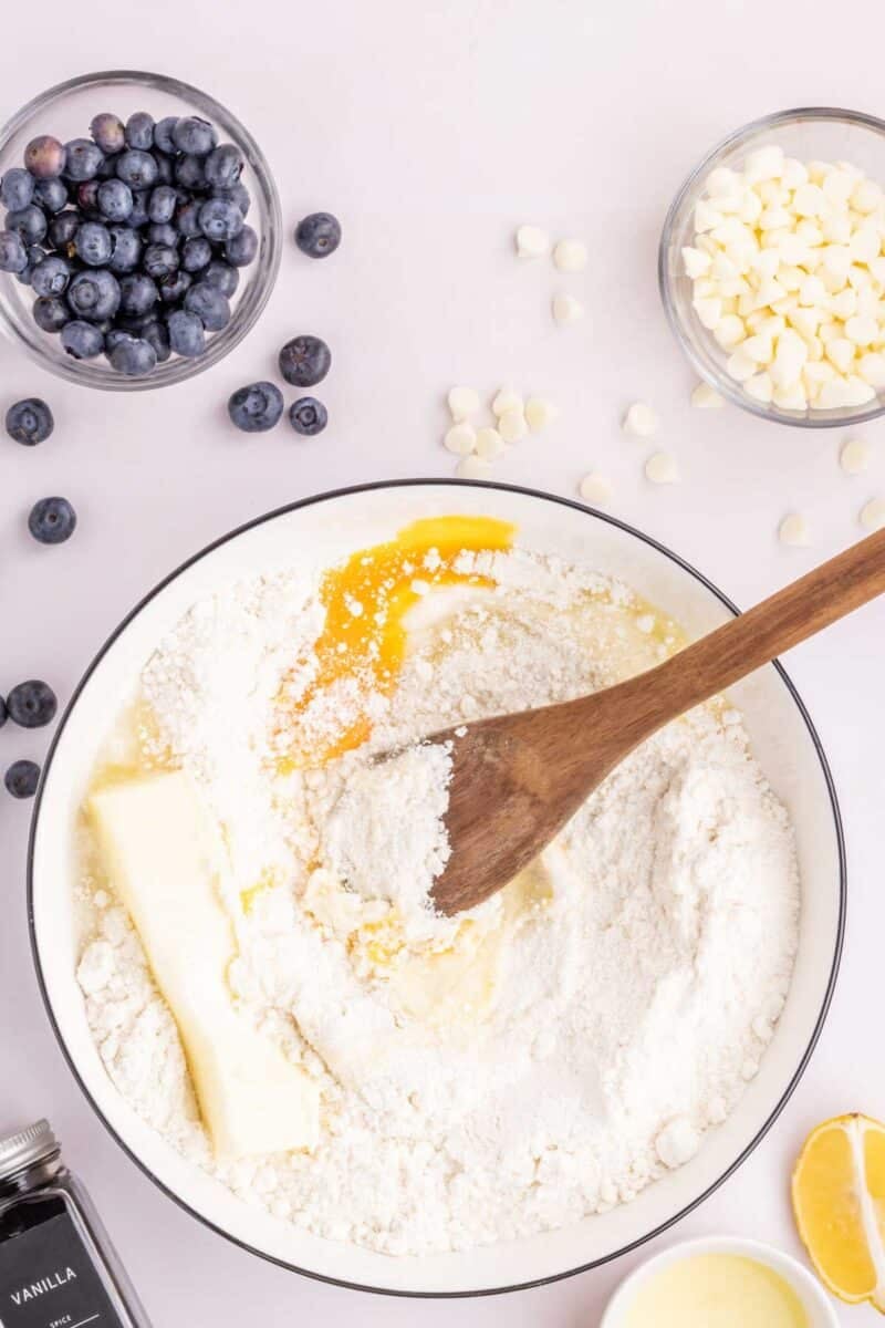 A mixing bowl with flour, butter, egg, and a wooden spoon, surrounded by bowls of blueberries, white chocolate chips, vanilla extract, and a lemon wedge.