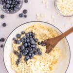 A mixing bowl with dough, fresh blueberries, and white chocolate chips being stirred with a wooden spoon, surrounded by small bowls of ingredients on a white surface.