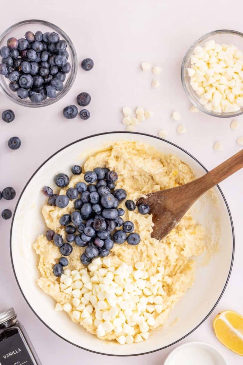A mixing bowl with dough, fresh blueberries, and white chocolate chips being stirred with a wooden spoon, surrounded by small bowls of ingredients on a white surface.