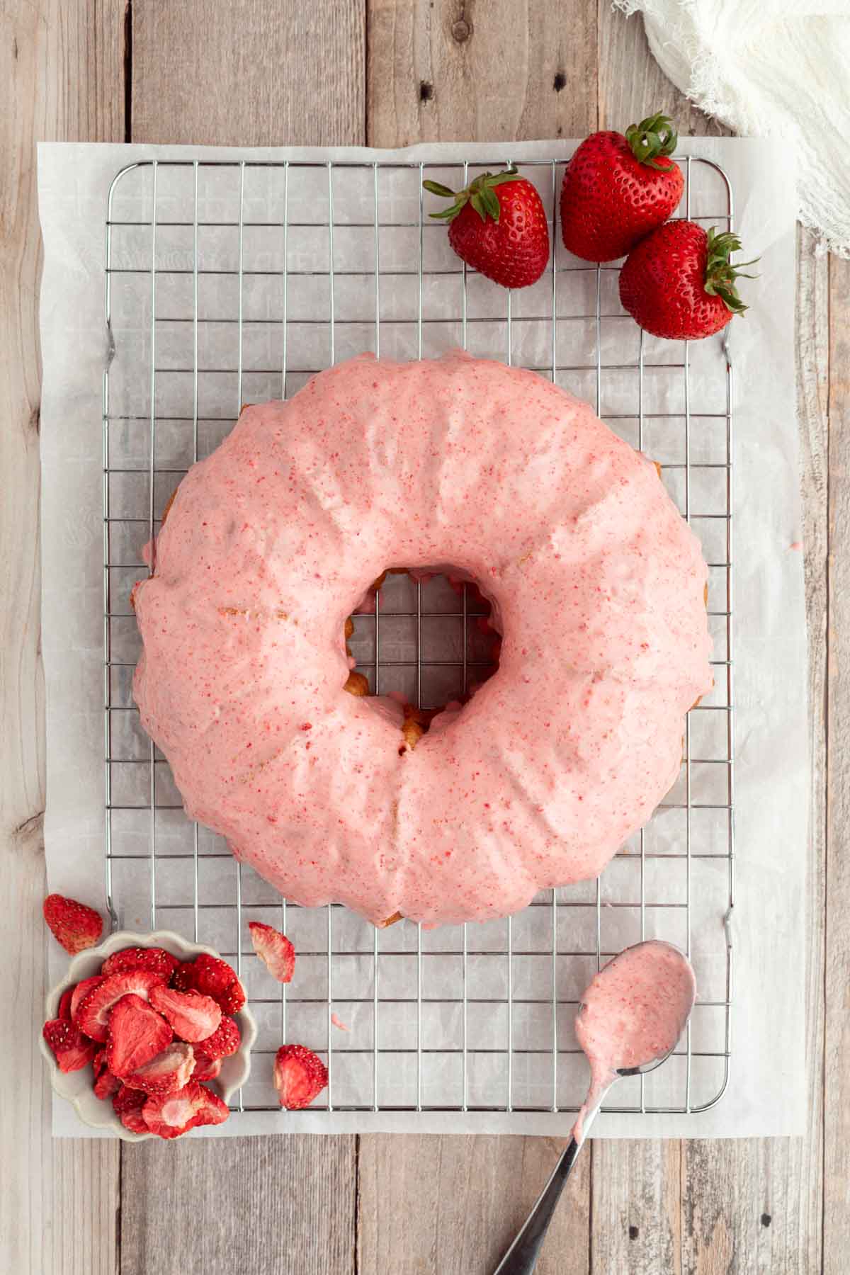A bundt cake with pink strawberry glaze sits on a cooling rack with fresh strawberries and a spoon beside it.