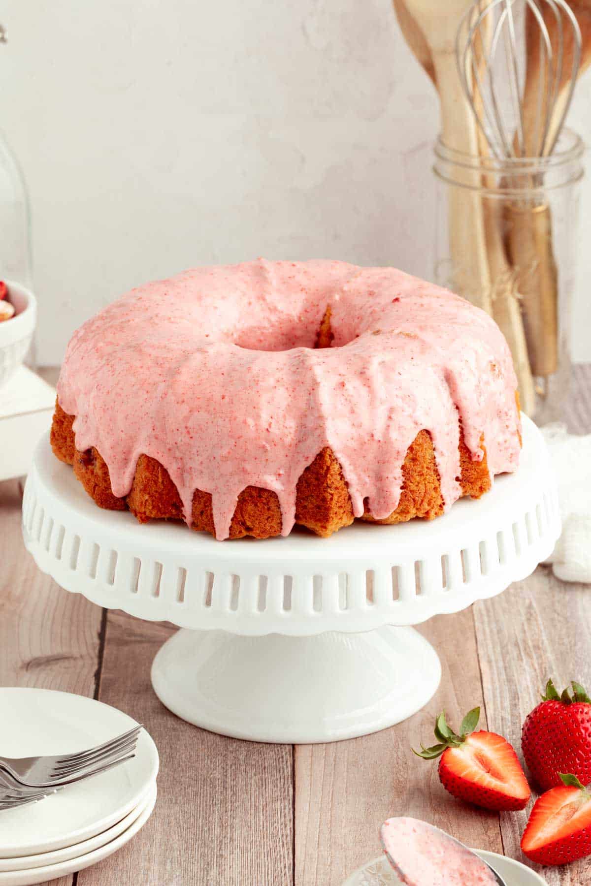A bundt cake with pink icing sits on a white cake stand, surrounded by fresh strawberries and kitchen utensils in the background.