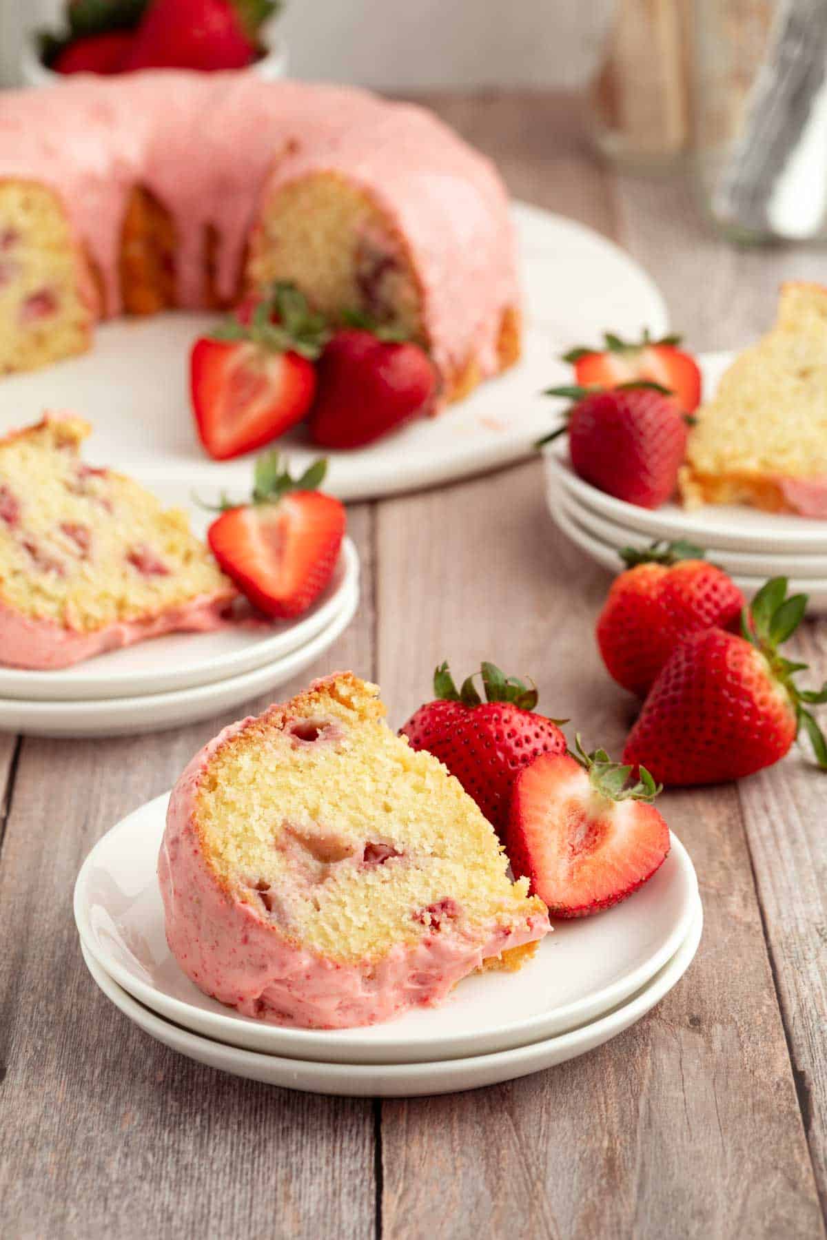 A slice of strawberry bundt cake with pink icing is served on a plate with fresh strawberries; the whole cake and more slices are in the background.