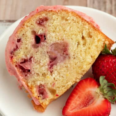 A slice of strawberry bundt cake with visible strawberry pieces, served on a white plate with two fresh strawberries beside it.