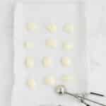 A parchment-lined baking sheet with twelve evenly spaced scoops of dough, plus a metal ice cream scoop resting at the bottom right.