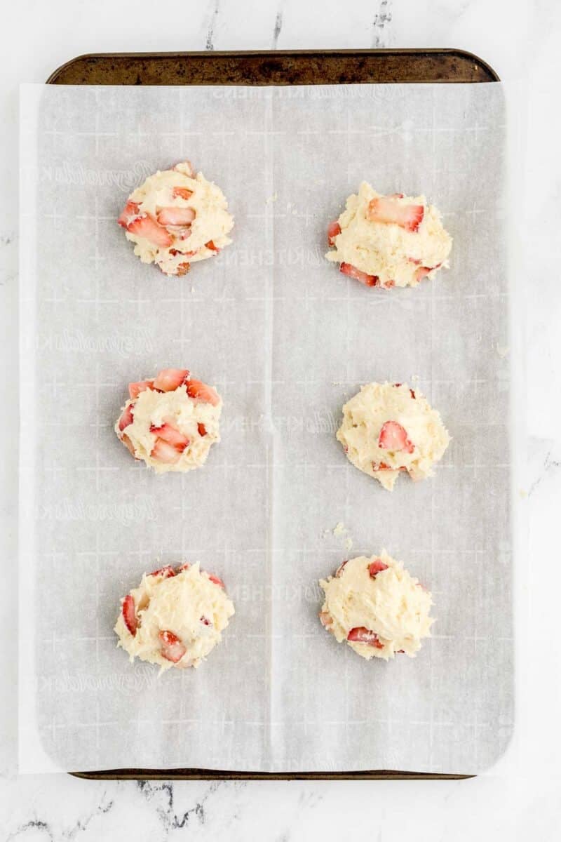 Six unbaked strawberry scones are arranged on a baking sheet lined with parchment paper, ready to go into the oven.