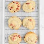 Six cookies with visible strawberry pieces are cooling on a wire rack placed on a white baking sheet over a marble surface.