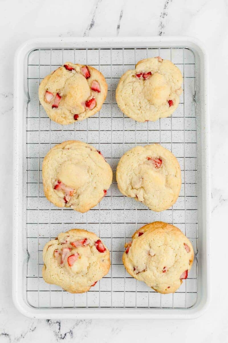 Six cookies with visible strawberry pieces are cooling on a wire rack placed on a white baking sheet over a marble surface.