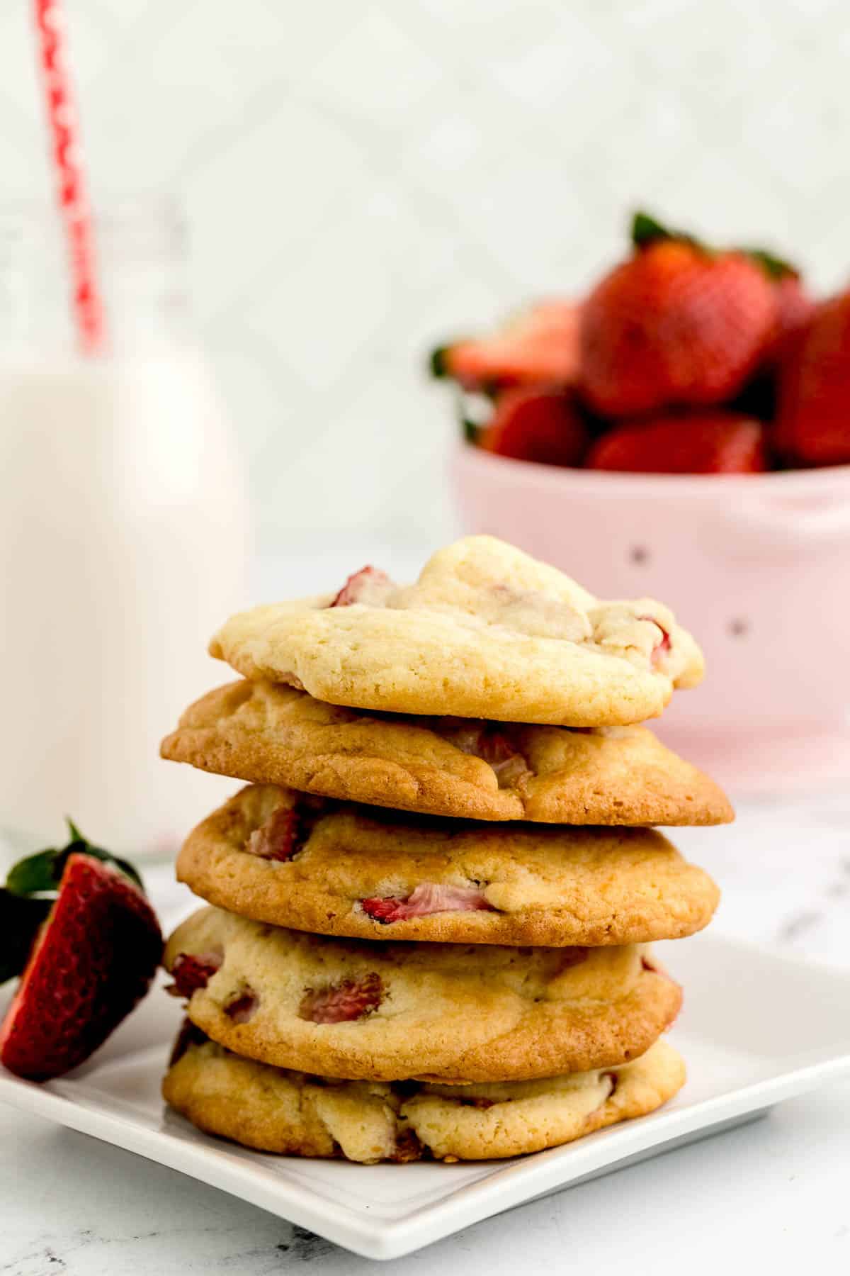 A stack of strawberry cookies on a white plate with a sliced strawberry, a bottle of milk, and a bowl of strawberries in the background.