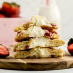 A stack of cookies filled with cream and strawberries sits on a wooden board, with fresh strawberries and a bowl of strawberries in the background.