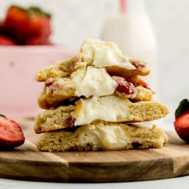 A stack of cookies filled with cream and strawberries sits on a wooden board, with fresh strawberries and a bowl of strawberries in the background.