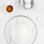 A glass bowl with sugar and butter, a small bowl with two eggs, and a small bowl of vanilla extract on a white marble surface.
