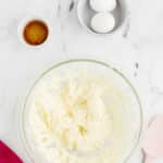 A glass bowl with creamed butter and sugar, a pink spatula, two eggs in a bowl, and a small dish of vanilla extract on a marble surface.