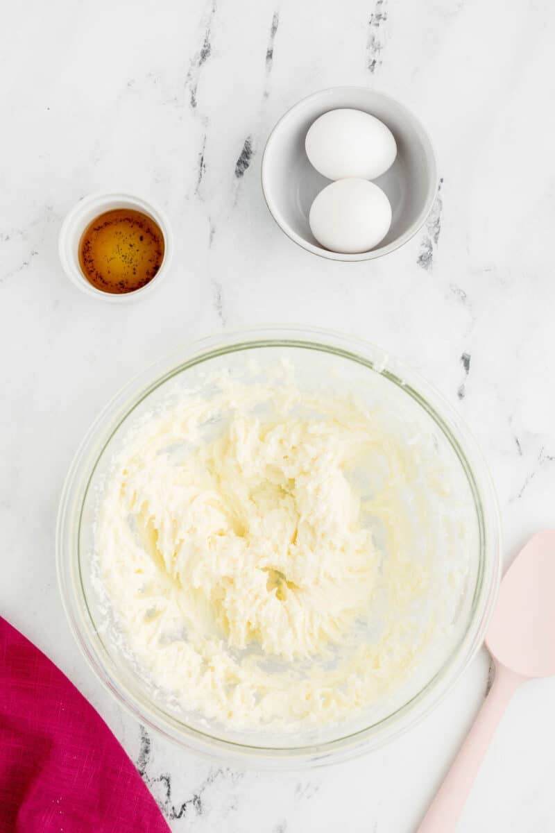 A glass bowl with creamed butter and sugar, a pink spatula, two eggs in a bowl, and a small dish of vanilla extract on a marble surface.