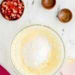 A glass bowl with creamed mixture and white powder, a bowl of chopped strawberries, two small empty bowls, and a pink spatula on a marble surface.