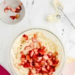 A glass bowl with cookie dough and chopped strawberries, a bowl with more strawberries, a hand mixer with dough residue, and a pink spatula on a marble surface.