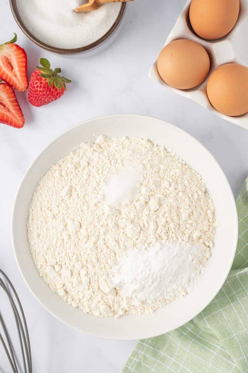 A bowl of flour with baking powder and baking soda sits on a counter next to eggs, sugar, strawberries, a whisk, and a green checkered cloth.