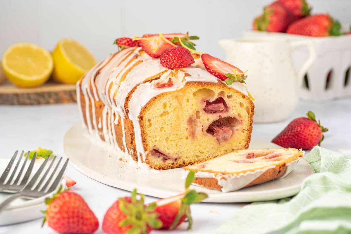 A loaf of strawberry lemon bread with white icing is sliced on a plate, surrounded by fresh strawberries and lemons.