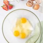 A glass bowl with three cracked eggs and sugar, surrounded by strawberries, a cup of yogurt, vanilla extract, broken eggshells, a whisk, and a green cloth on a white surface.