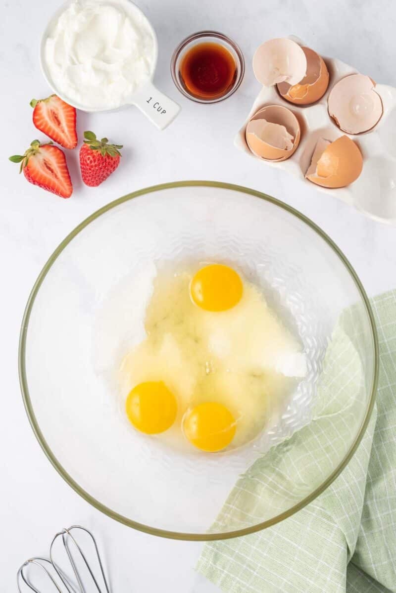 A glass bowl with three cracked eggs and sugar, surrounded by strawberries, a cup of yogurt, vanilla extract, broken eggshells, a whisk, and a green cloth on a white surface.