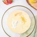 A glass bowl with a yellow batter mixture, lemon zest, and yogurt on top, surrounded by strawberries, a lemon slice, a mixer, and a green kitchen towel.