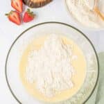 A glass bowl with flour on top of a yellow batter, next to a bowl of flour, sliced strawberries, a lemon half, and a wooden citrus reamer on a cutting board.