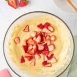 A glass bowl filled with yellow cake batter and chopped strawberries on top, surrounded by a pink spatula, fresh strawberries, a lemon, and a wooden spoon on a plate.