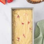 A loaf pan filled with cake batter containing pieces of strawberries, with fresh strawberries and a halved lemon beside it on a marble surface.