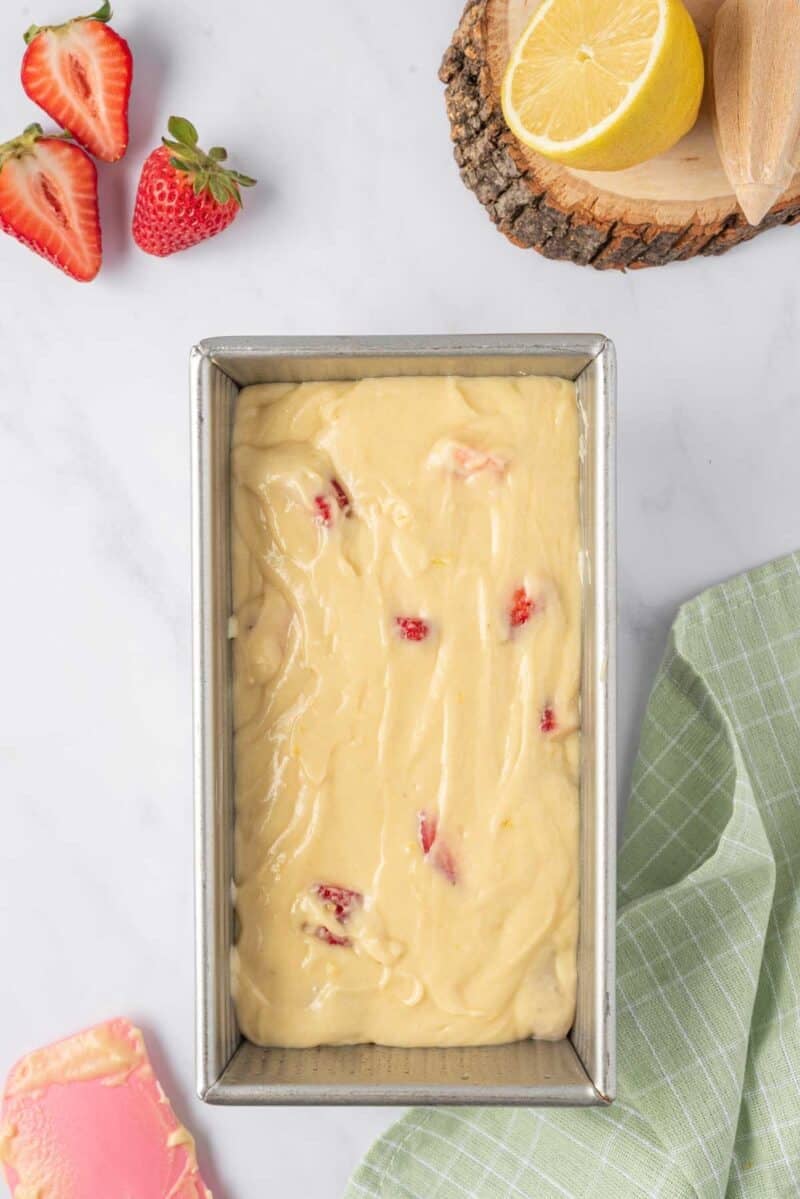 A loaf pan filled with cake batter containing pieces of strawberries, with fresh strawberries and a halved lemon beside it on a marble surface.