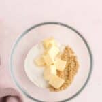 A glass bowl containing cubed butter, granulated sugar, and brown sugar on a light pink surface.