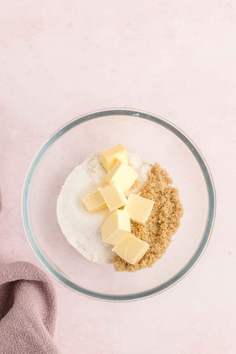 A glass bowl containing cubed butter, granulated sugar, and brown sugar on a light pink surface.