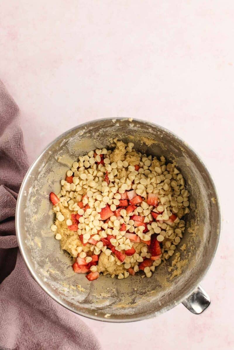 A mixing bowl containing cookie dough with white chocolate chips and chopped strawberries on a light pink surface.