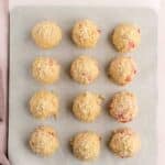Twelve unbaked cookie dough balls with visible red bits and crumb topping are arranged on a parchment-lined baking sheet.
