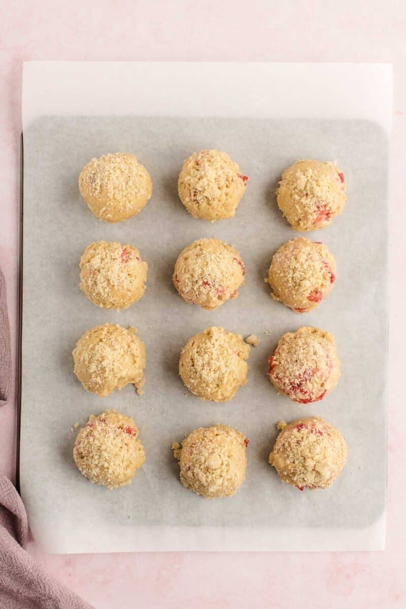 Twelve unbaked cookie dough balls with visible red bits and crumb topping are arranged on a parchment-lined baking sheet.