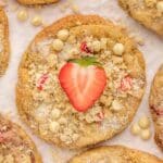 Close-up of strawberry shortcake cookies topped with white chocolate chips, crumbly streusel, and half a fresh strawberry, arranged on a white surface.