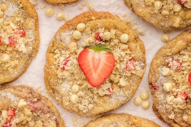Close-up of strawberry shortcake cookies topped with white chocolate chips, crumbly streusel, and half a fresh strawberry, arranged on a white surface.