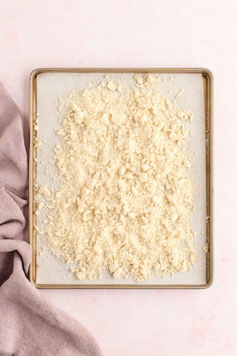 A baking tray lined with parchment paper holds an even layer of crumbled dough or streusel, with a mauve kitchen towel to the left.
