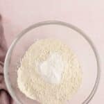 A clear glass bowl filled with flour and baking powder sits on a pink surface next to a folded mauve cloth.