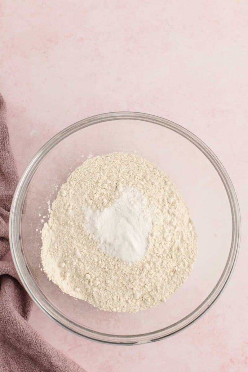 A clear glass bowl filled with flour and baking powder sits on a pink surface next to a folded mauve cloth.