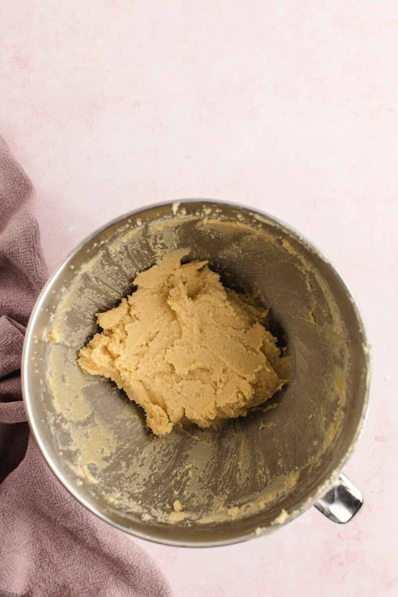 A metal mixing bowl contains a ball of cookie dough, placed on a light pink surface with a purple cloth partially visible on the side.