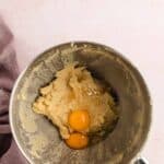 A metal mixing bowl containing creamed cookie dough and two raw eggs, viewed from above, with a purple cloth partially visible on the side.