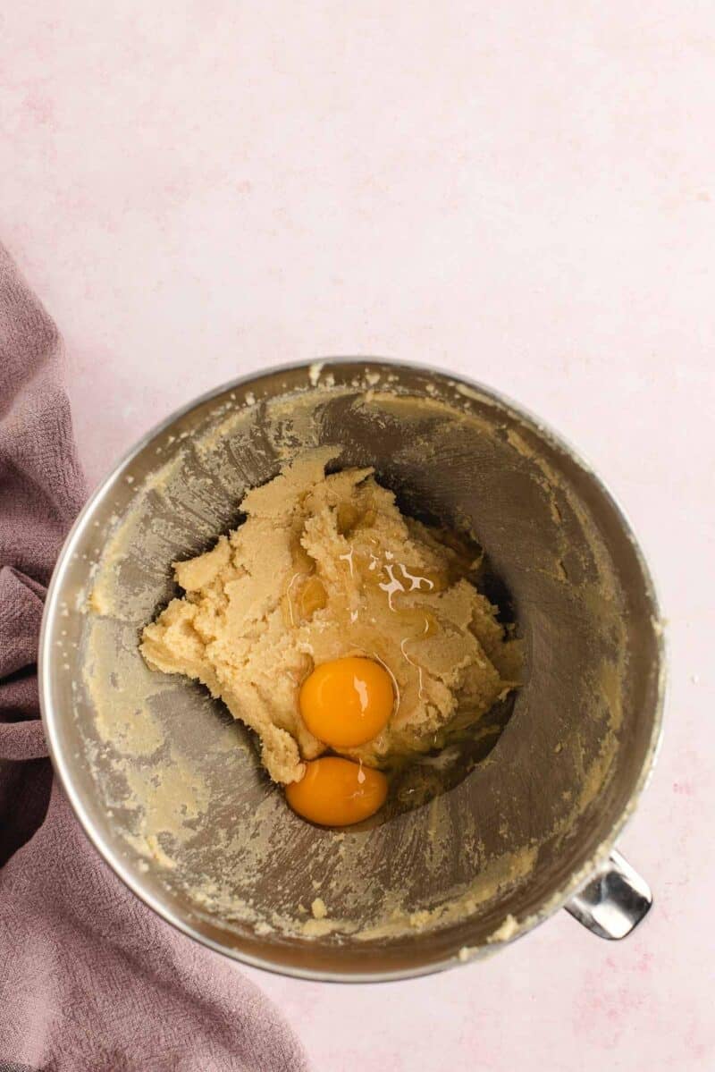 A metal mixing bowl containing creamed cookie dough and two raw eggs, viewed from above, with a purple cloth partially visible on the side.